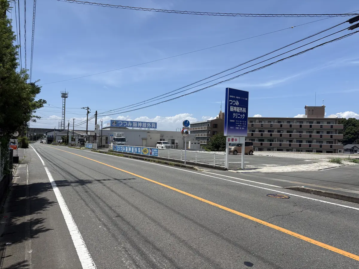 県道15号から見たつつみ脳神経外科クリニック。道路の右手に、青地に白抜きで「つつみ脳神経外科」と書かれた横長の建物看板と、診療時間を記した縦長の案内看板が並んで立っている。手前は白いガードフェンスで、その奥が駐車場。