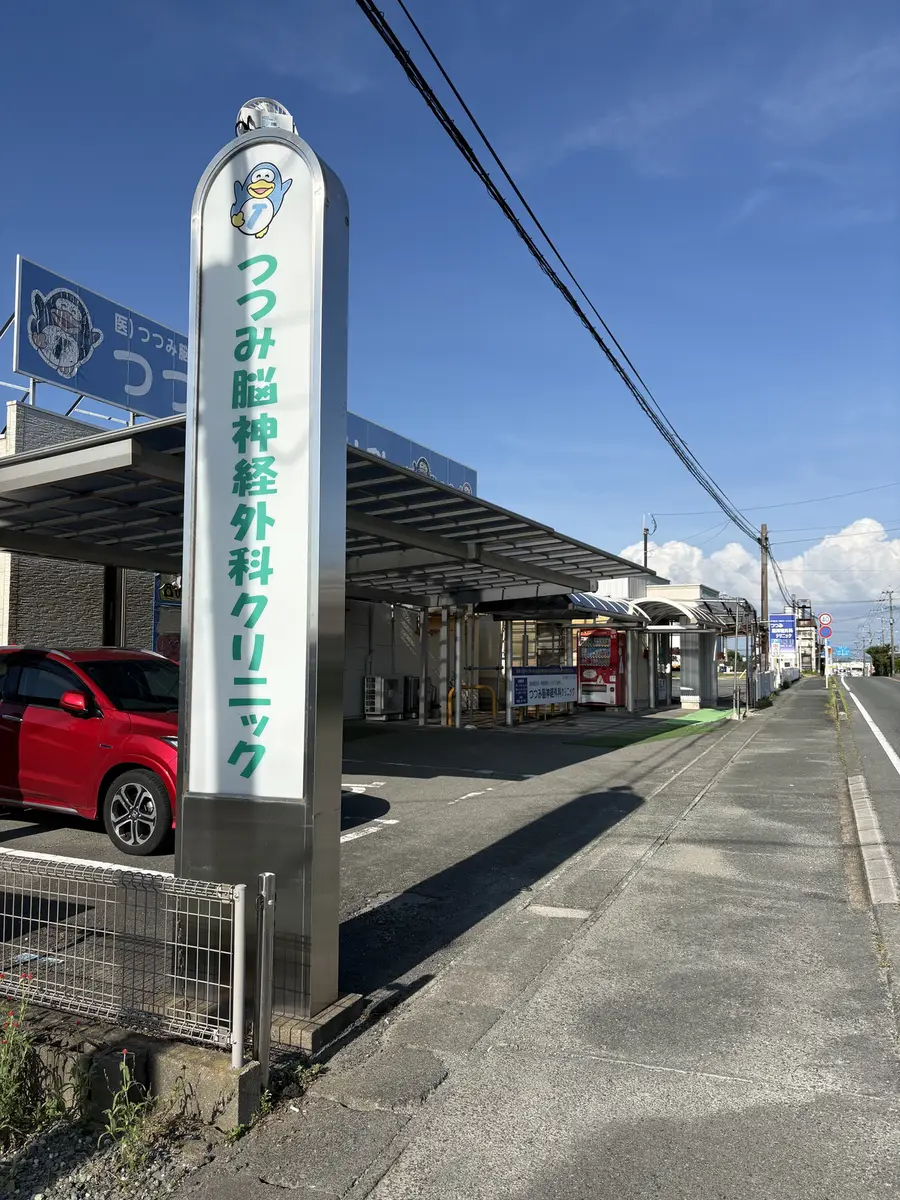 つつみ脳神経外科クリニックの縦長の案内看板。上部に青いペンギンのマークがあり、その下に「つつみ脳神経外科クリニック」と緑色の文字で縦書きされている。奥には屋根付きのスペースが続いている。