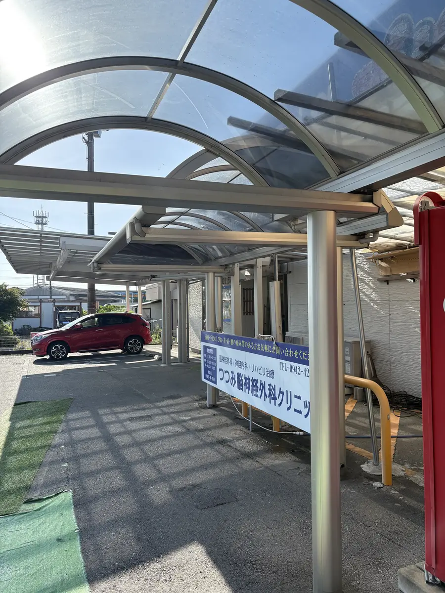 クリニックの入口まわりに架かるアーチ状の透明な屋根。屋根の下は通路になっており、「つつみ脳神経外科クリニック 脳神経外科／神経内科／リハビリ治療」と書かれた青い看板が掲げられている。奥には屋根のない駐車場と停まっている車が見える。
