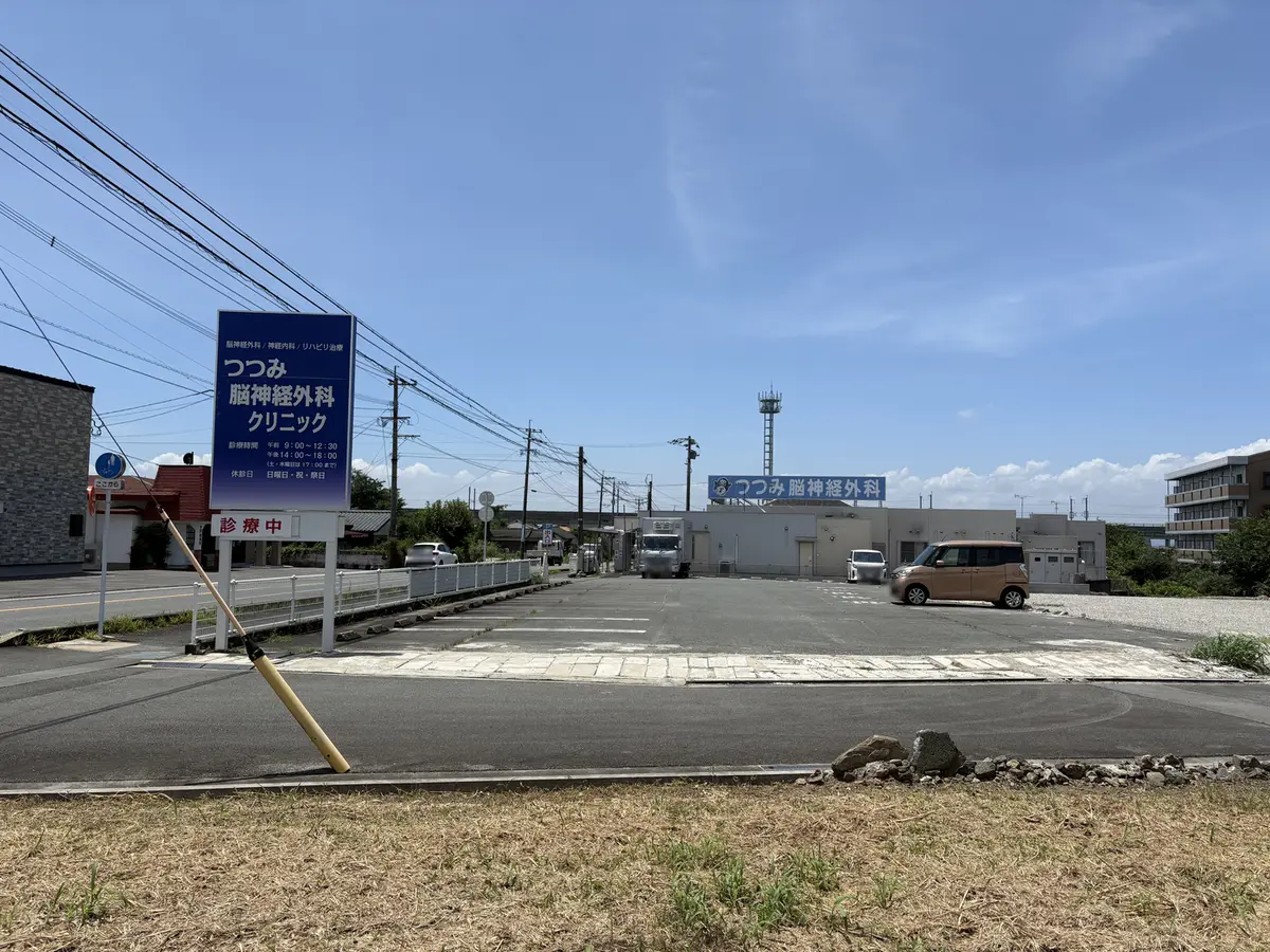 つつみ脳神経外科クリニックの駐車場。白線で区切られた広い平面駐車場で、多くの区画が空いている。奥にクリニックの建物と「つつみ脳神経外科」の看板が見える。
